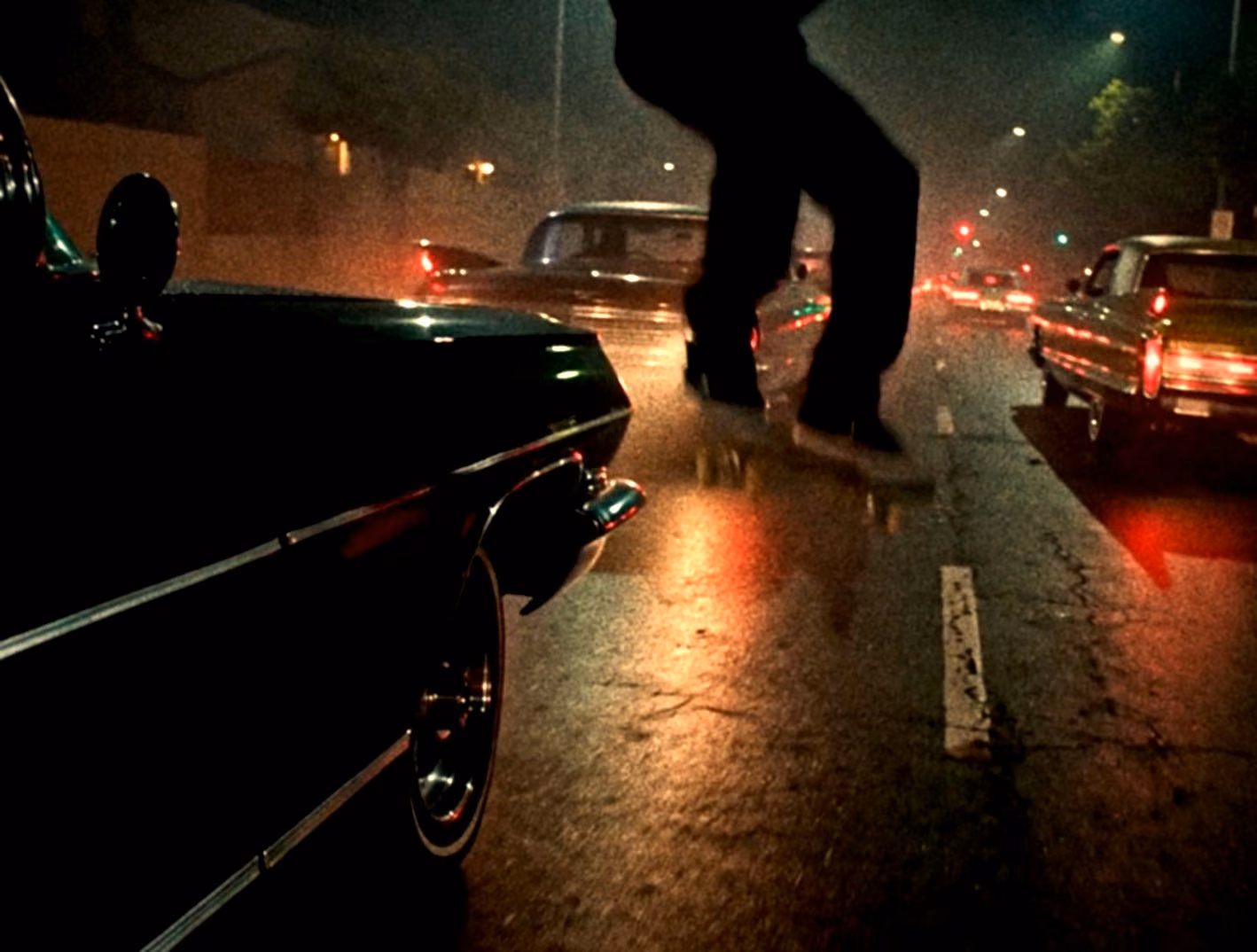 A figure jumping between lowriders on a wet city street